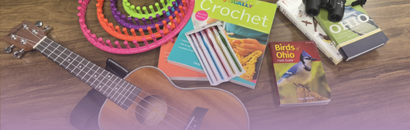 Library of Things items arranged on a wooden table, including a ukulele, colorful knitting looms, a crochet book and hook set, a Birds of Ohio field guide, binoculars, and an Ohio guidebook.