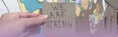 A hand holds a handwritten "We Are Hiring" post it note in front of a colorful painted mural.